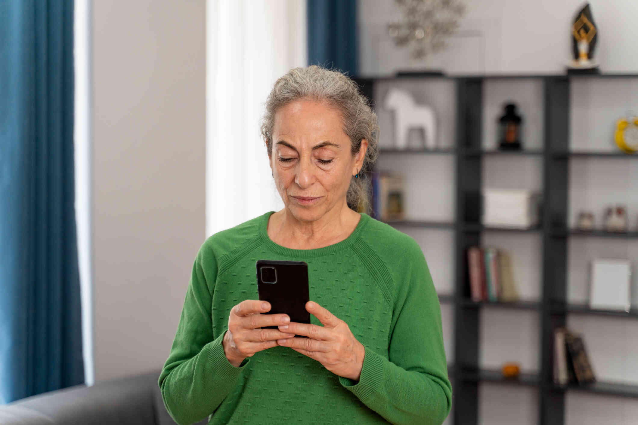 A mature woman in a green sweater stands in her home and looks down at the phone in her hand.